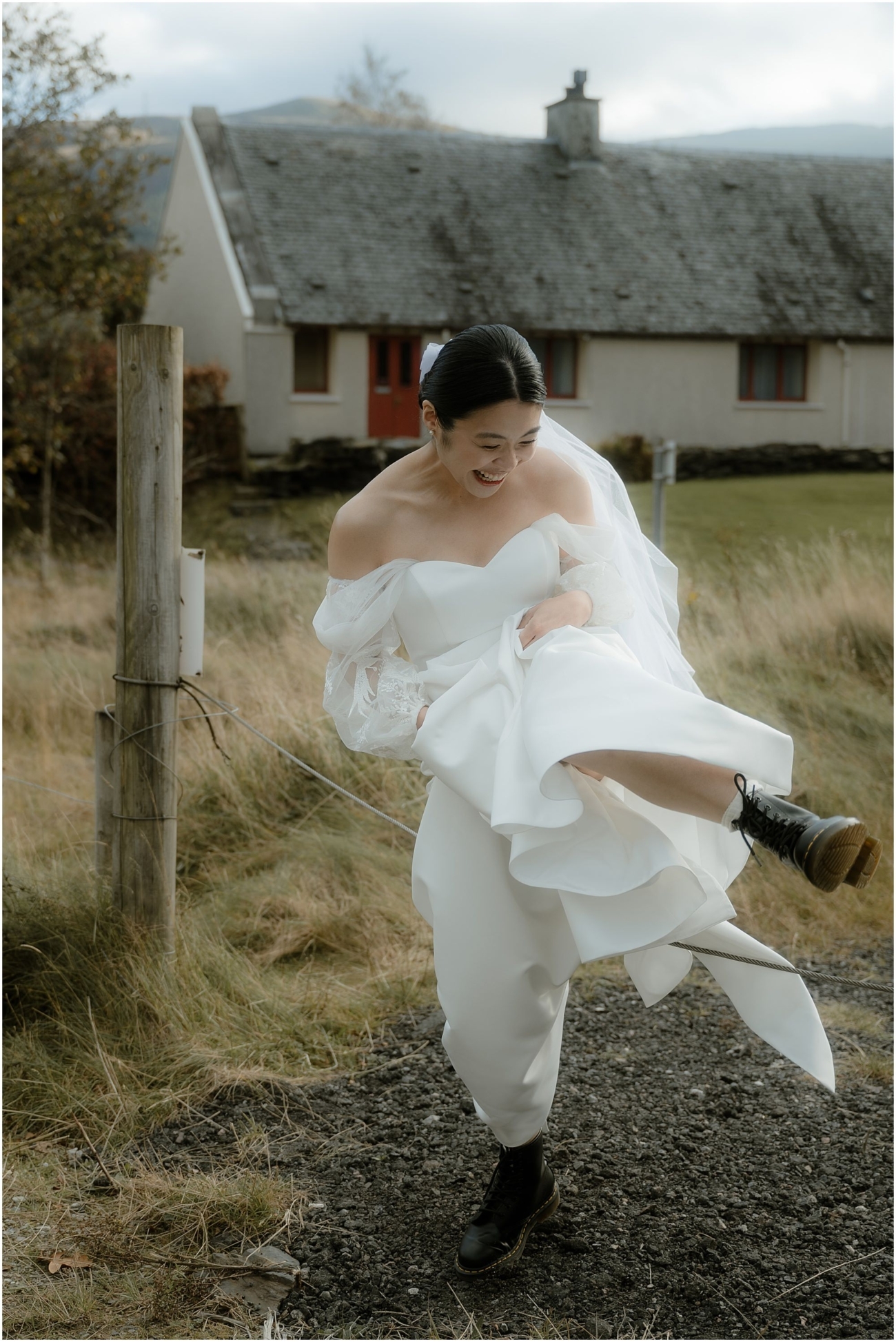 Bride playfully lifting her dress and laughing as she steps over a fence wire in her wedding boots