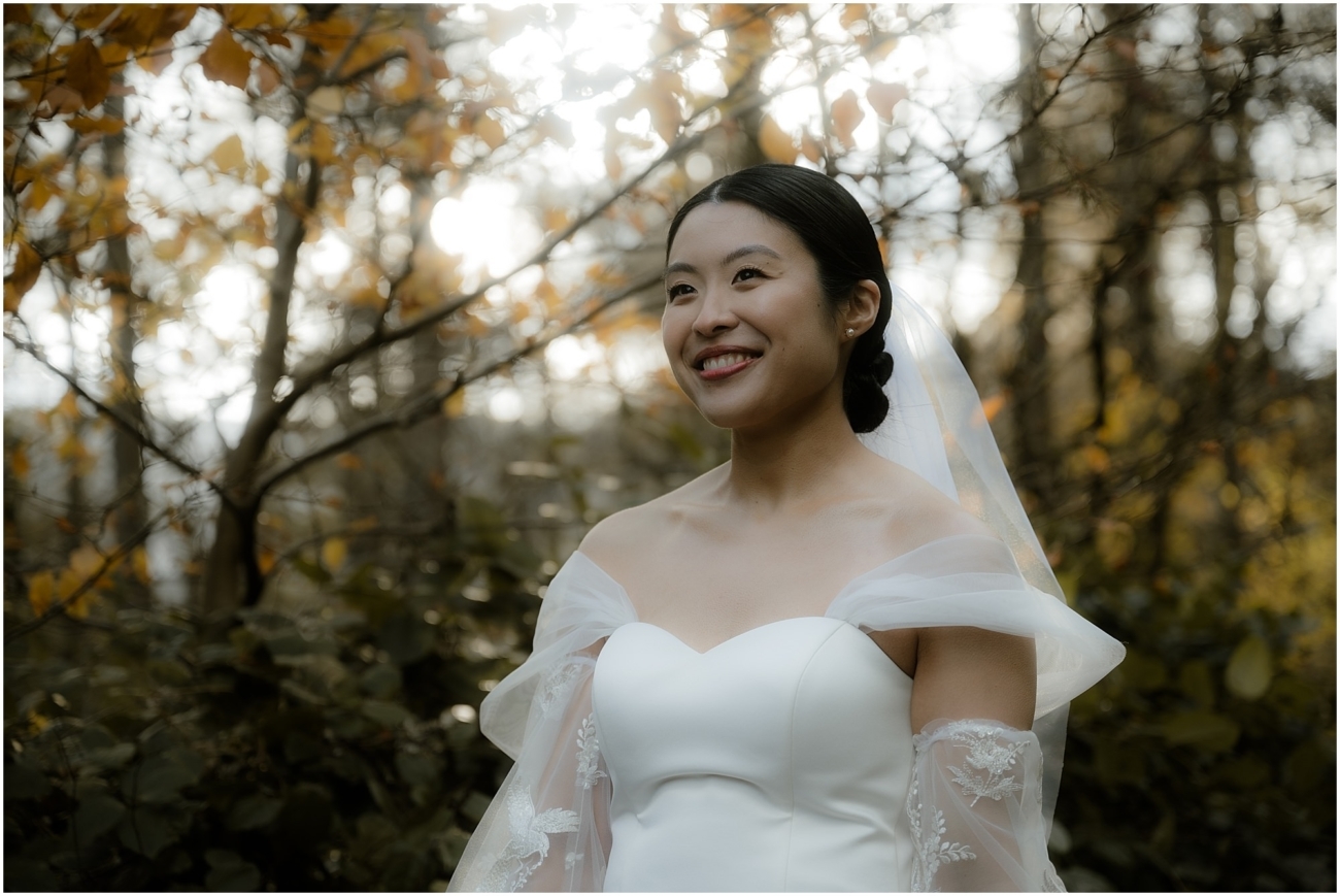 Bride stands in a woodland setting with golden autumn leaves, smiling as sunlight filters through the branches