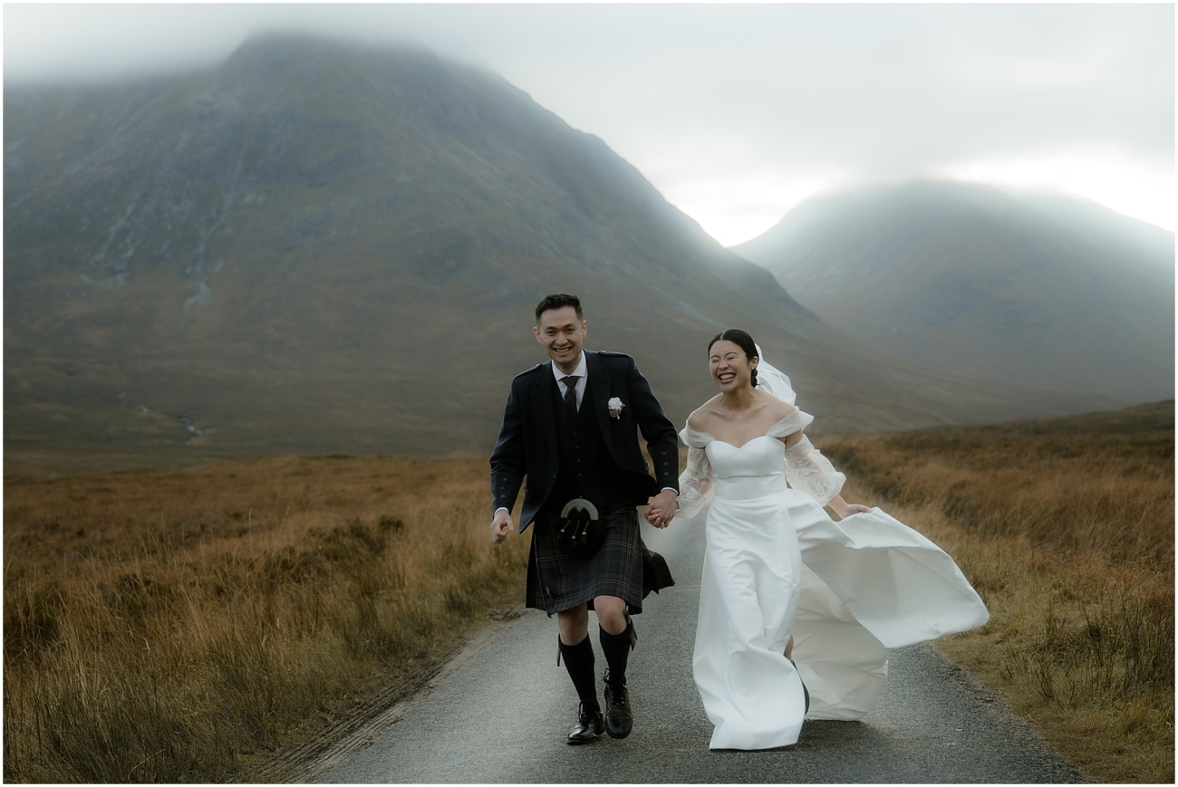 The couple runs hand in hand down a road in the Highlands, with cloud-covered mountains in the background