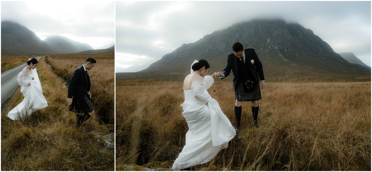 The groom helps the bride step off the road into a grassy field, surrounded by towering mountains