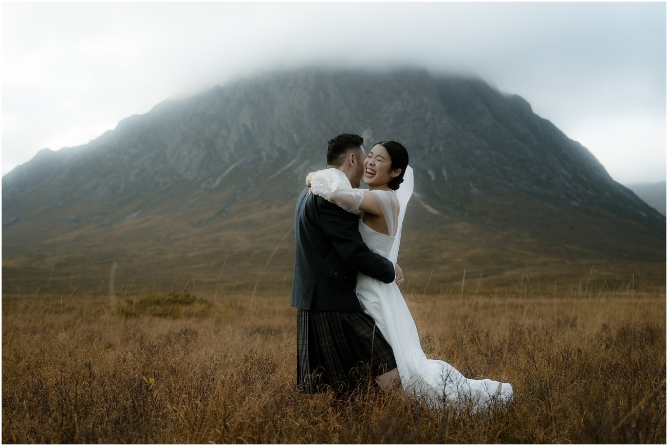 The bride and groom embrace in an open field in front of a mountain, laughing