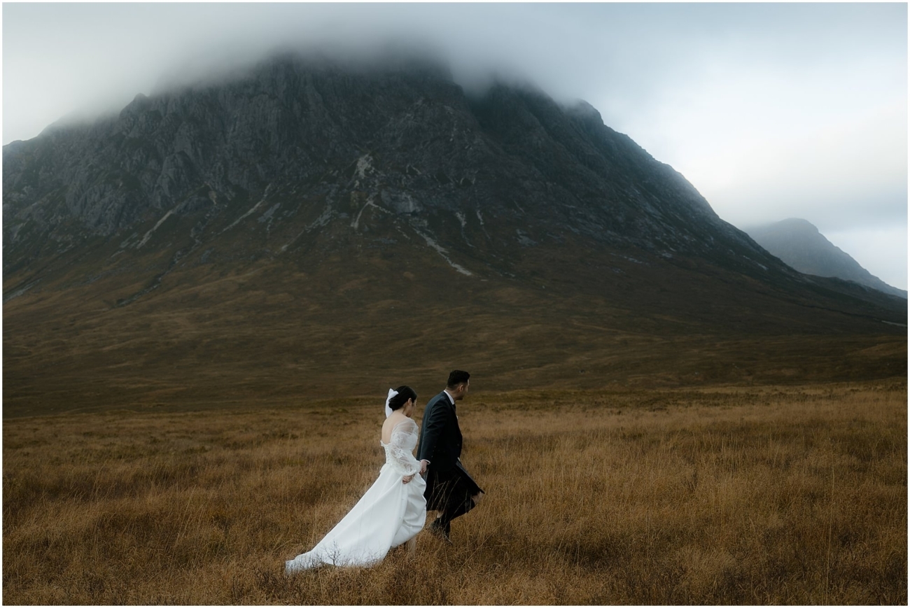 The couple walks side by side through a golden field, the bride's dress trailing behind her, with the mountain looming above them