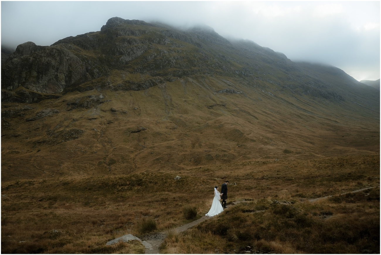 A small figure of a couple walking along a narrow trail in the Scottish Highlands, dwarfed by a rugged mountain landscape