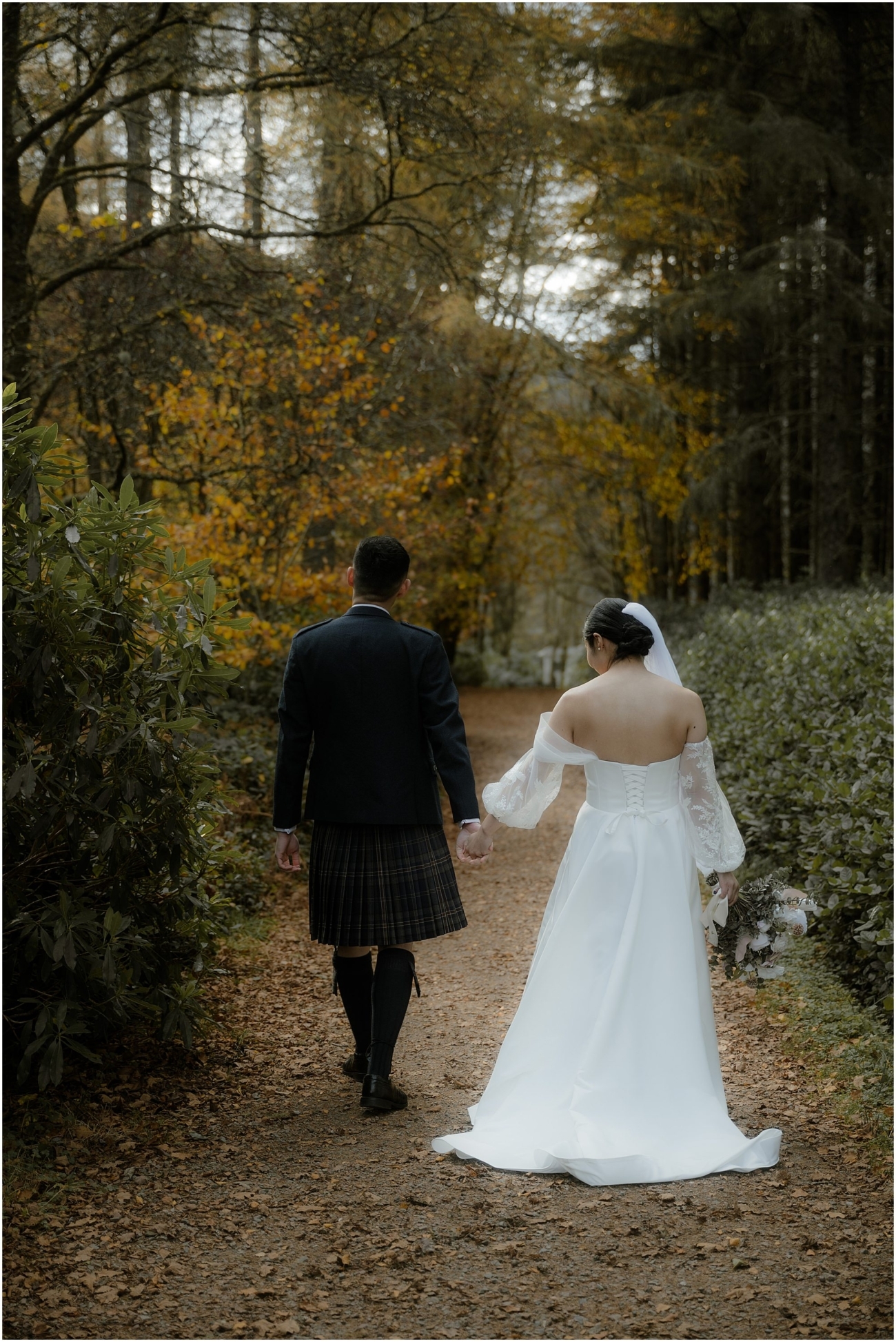 Bride and groom walking hand in hand along a forest path with golden autumn leaves