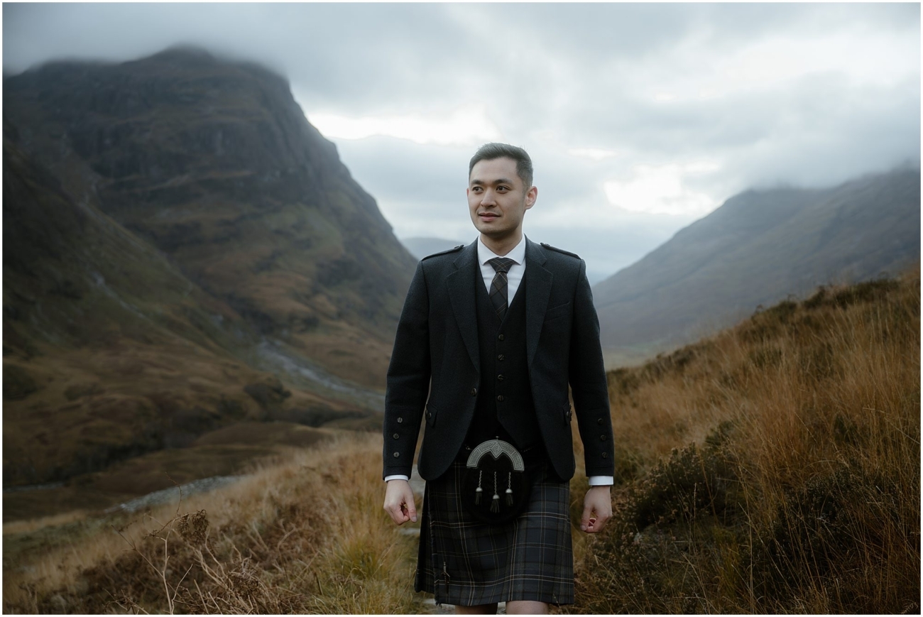 A groom in a traditional Scottish kilt walking down a narrow hiking trail