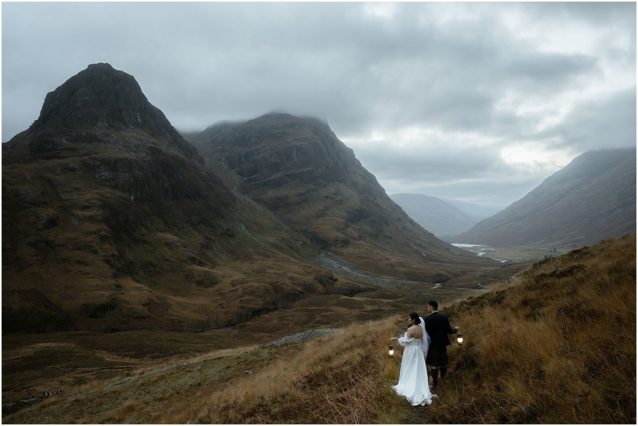 A wide shot of the couple walking along a Highland trail, holding lanterns, with sweeping mountain views