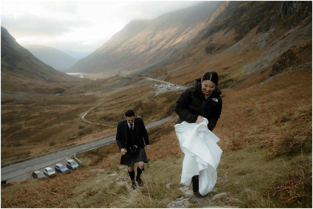 The bride hiking up a steep Highland trail, bundled in a black puffer jacket over her wedding dress, as the groom in a kilt follows behind her