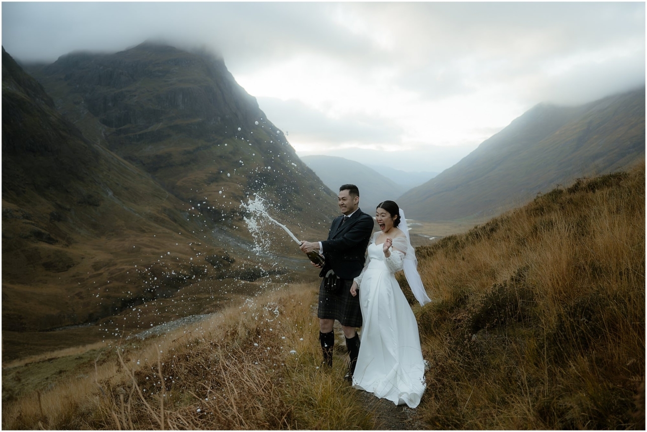 The couple celebrating in the Scottish mountains, popping open a bottle of champagne, with bubbles catching the light against the moody landscape