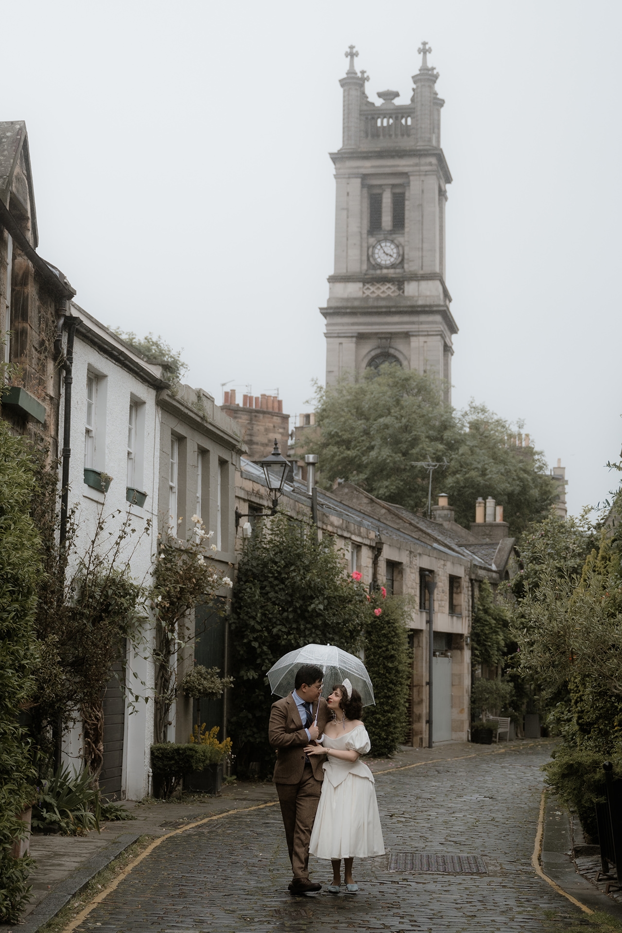 Couple walking under an umbrella on a rainy cobblestone street in Edinburgh, dressed in vintage-inspired elopement attire with a historic clock tower in the background