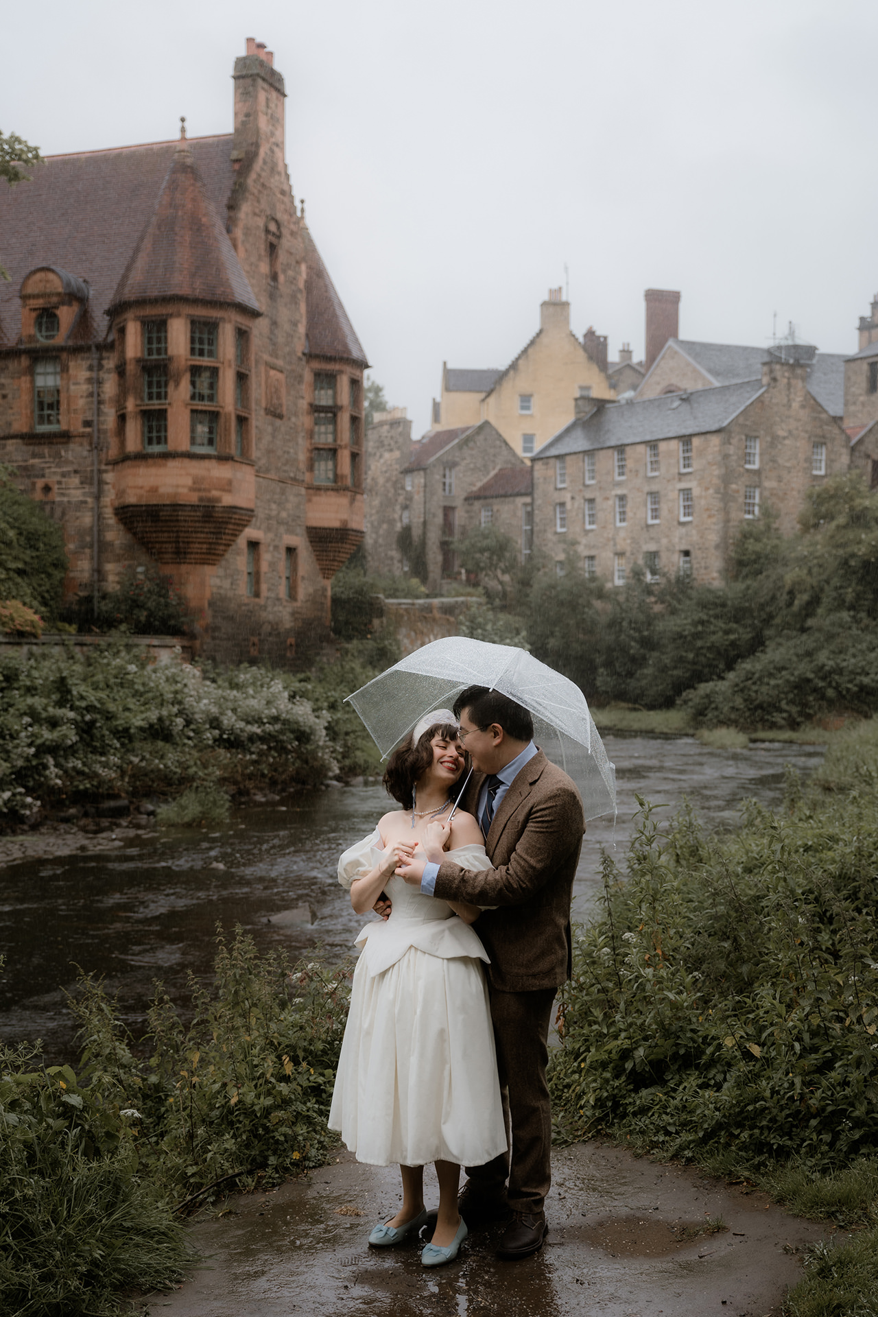 Romantic elopement couple embracing under an umbrella by the riverside in Dean Village, Edinburgh, surrounded by historic stone buildings and soft rain