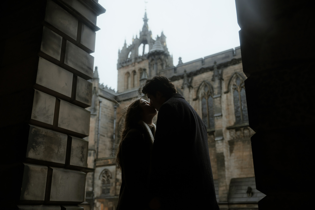 Dark academia-inspired engagement photoshoot on a rainy day in Edinburgh - bride and groom sharing a kiss in one of the arches behind St Giles Cathedral