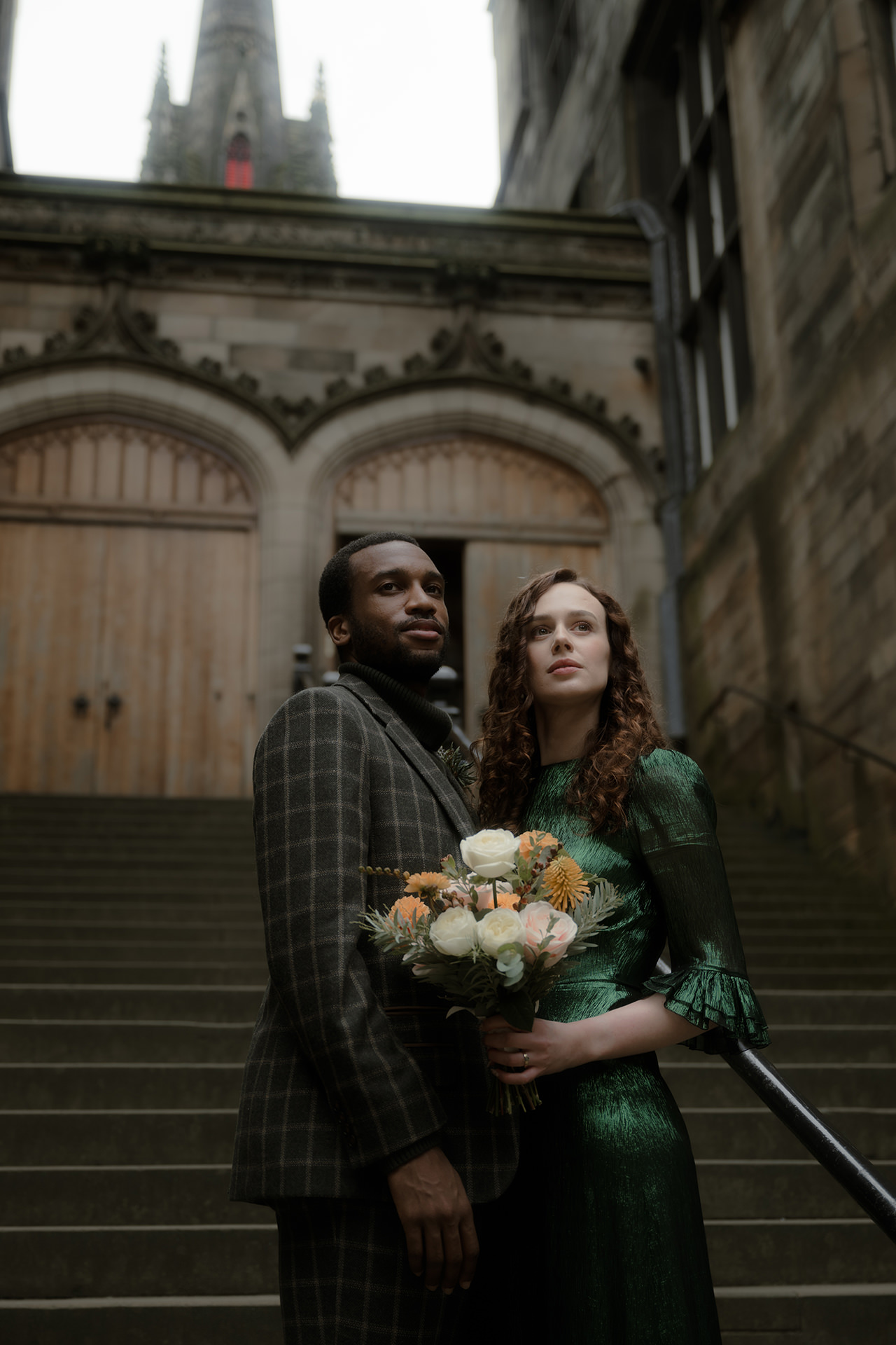 Stylish newlywed couple standing on the steps of University of Edinburgh New College, her holding a wedding bouquet 