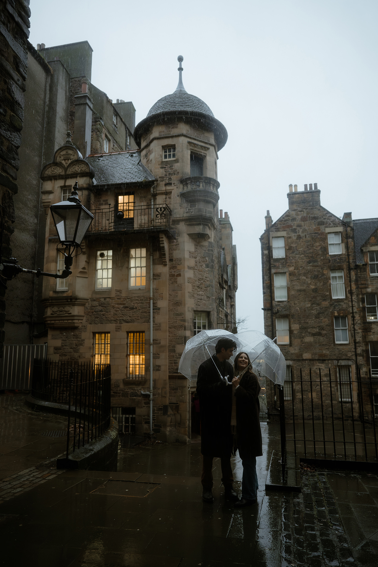 Engaged couple standing underneath clear umbrellas in front of the Writer's Museum in Edinburgh