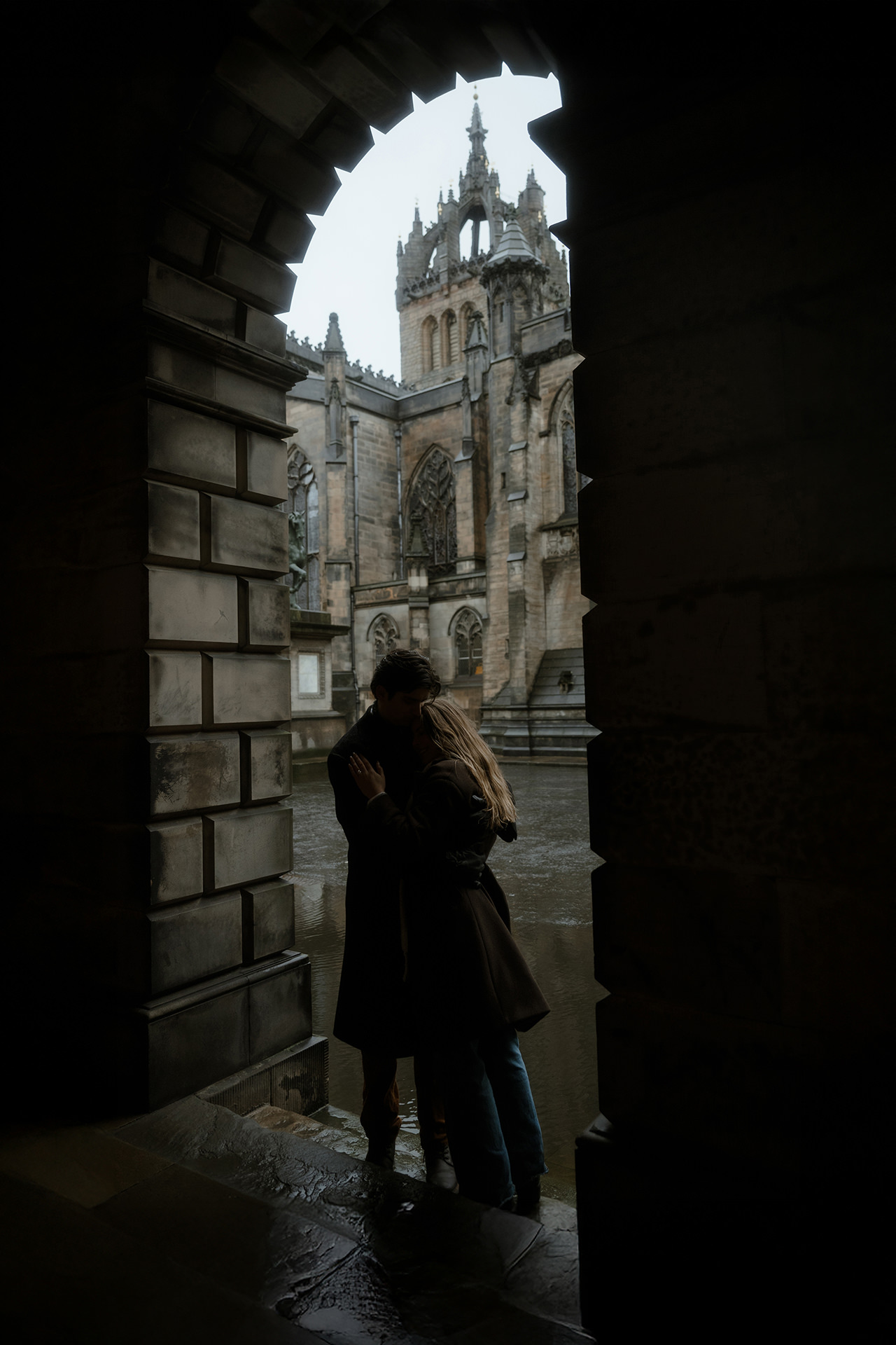 Couple sharing a close embrace under Royal Mile arches on a rainy Scottish day