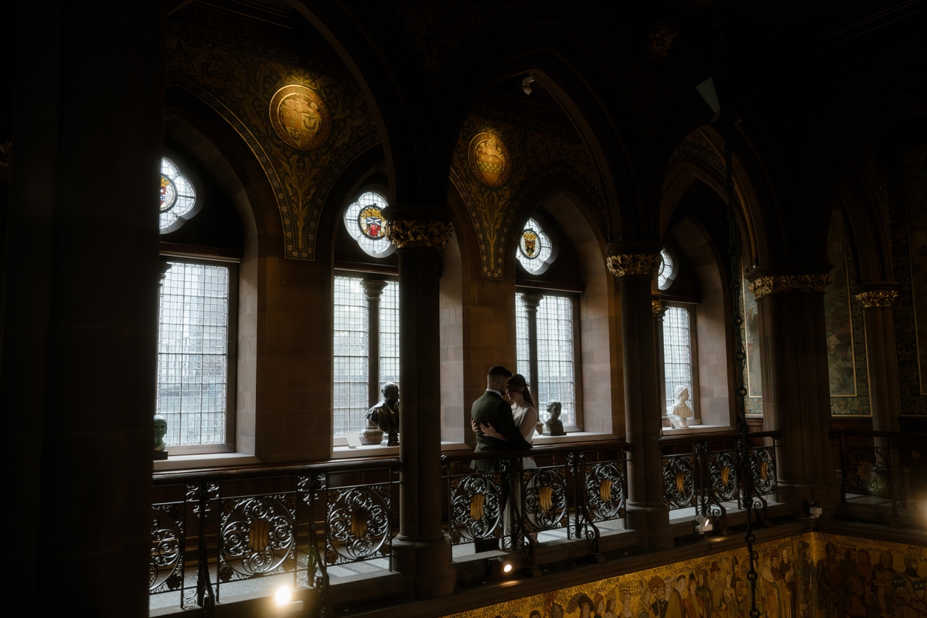 Bride in a simple white gown and groom in a dark suit embrace an ornate iron-and-stone gallery balcony inside a vaulted hall, framed by stained-glass medallion windows with classical bust sculptures on the sill, on their wedding day