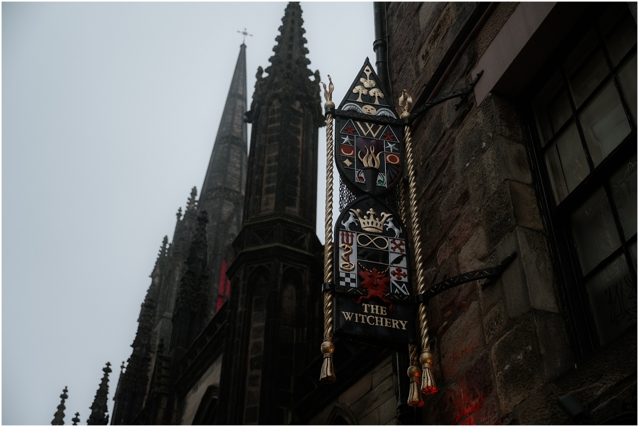 Exterior sign of The Witchery by the Castle in Edinburgh’s Old Town, with gothic spires and moody winter sky in the background.