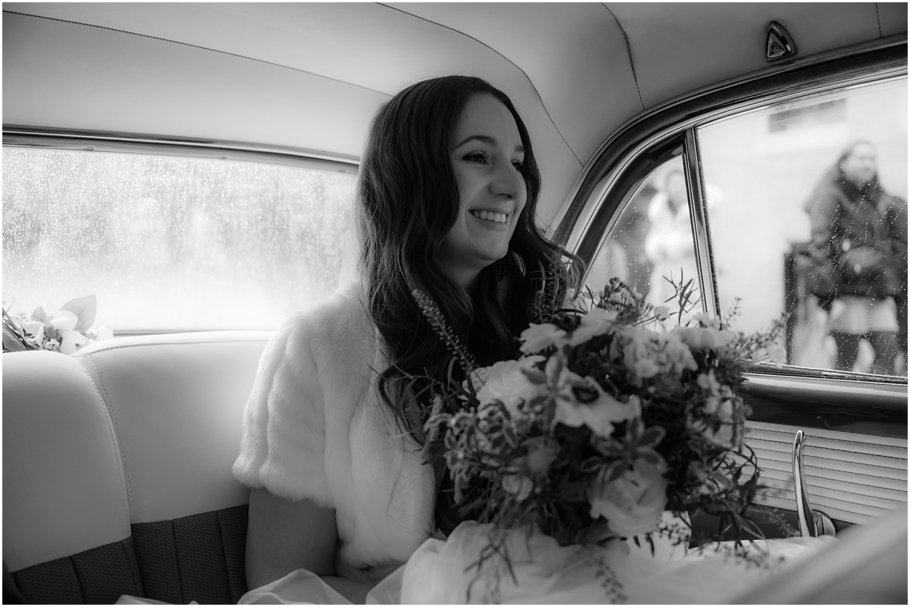 Black and white portrait of the bride seated inside a vintage wedding car, gazing softly through a rain-speckled window