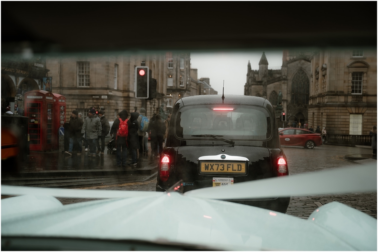 View through a classic wedding car windscreen of a rainy Edinburgh street scene, featuring a black London taxi in front, a red phone box, and crowds crossing near St Giles' Cathedral on the Royal Mile.