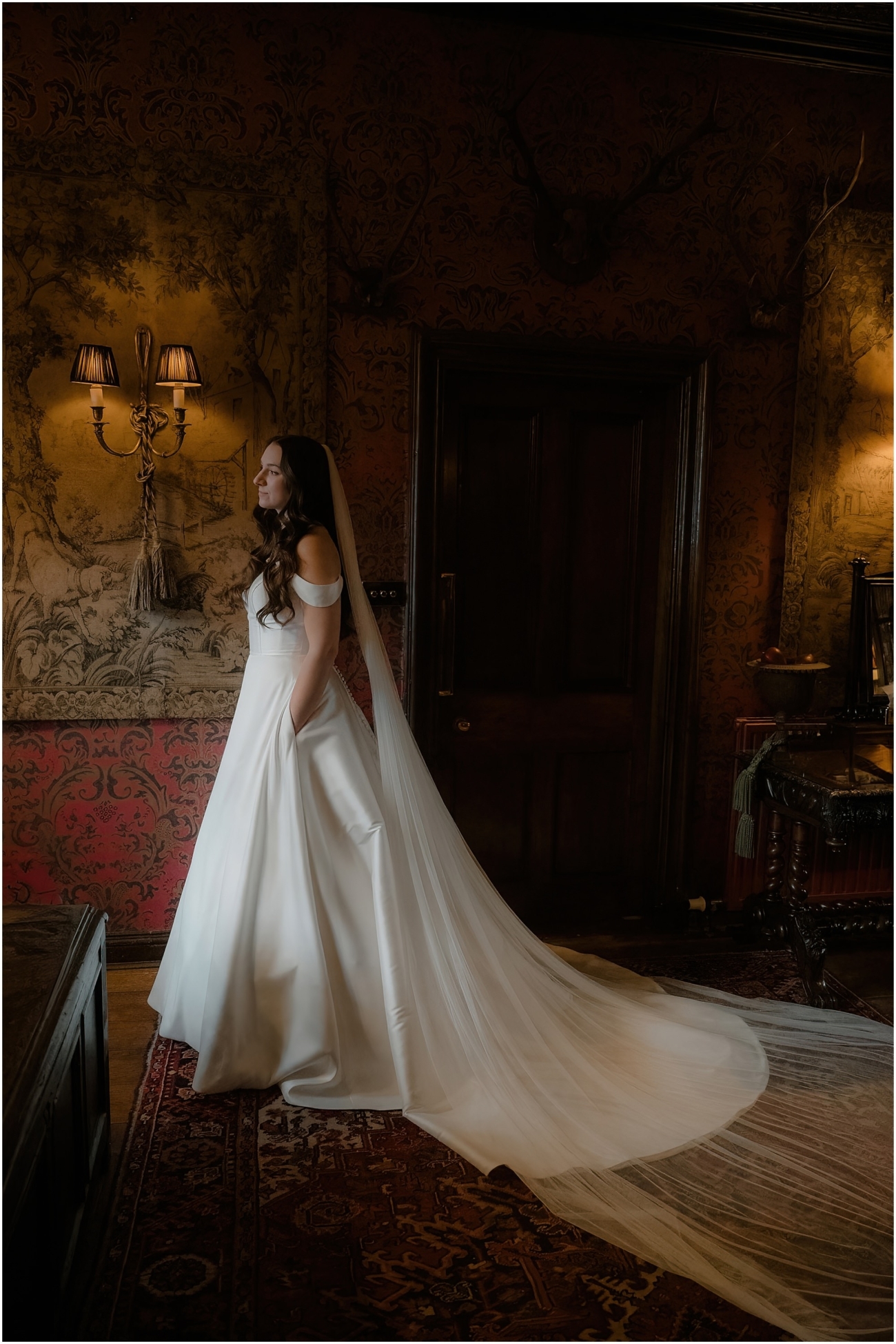 Full-length portrait of the bride in an off-shoulder wedding gown with long veil, posing beside a dramatic gothic fireplace and gold-framed portrait.