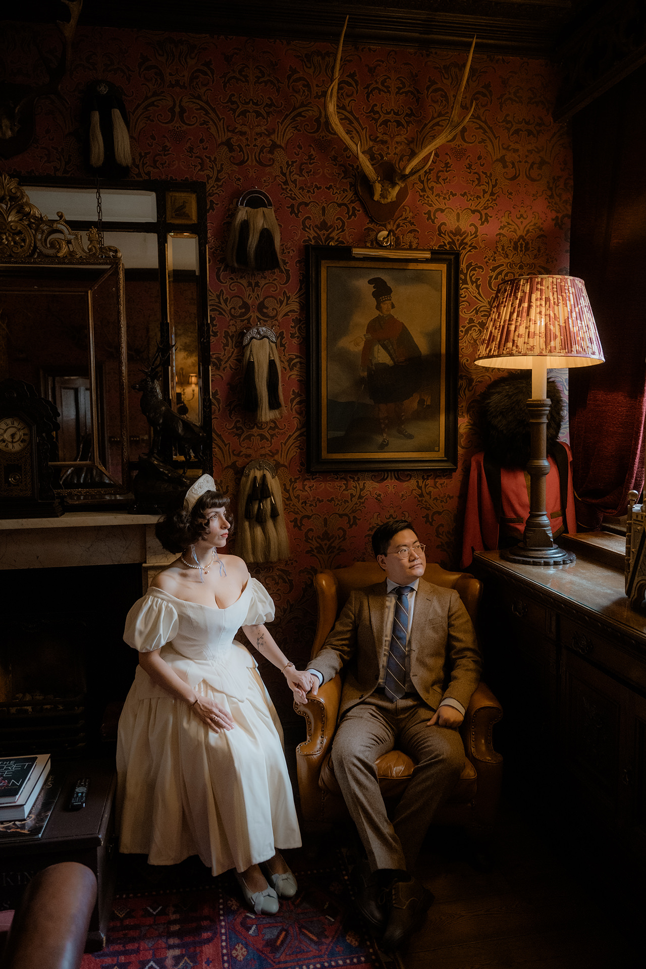 Bride and groom sitting side by side in their Witchery suite in Edinburgh with deer antlers and an old painting on the wall behind them