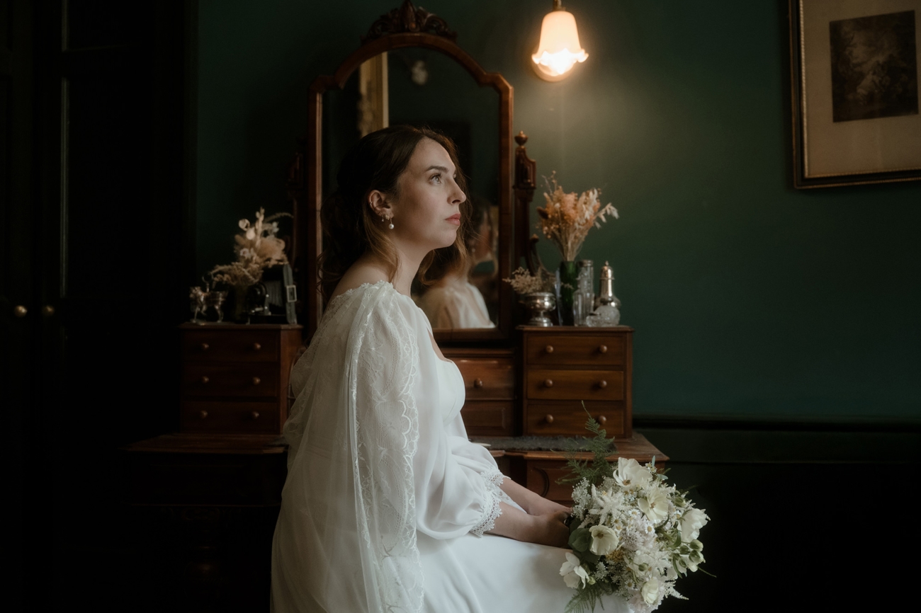 Romantic bridal portrait—bride in vintage lace-sleeved gown holding ivory bouquet, seated at antique vanity in dark green Victorian room