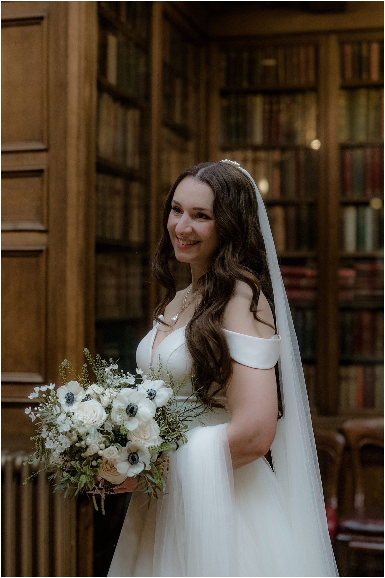 Bride holding a bouquet of white anemones and roses, smiling in front of antique bookcases at the Royal College of Physicians in Edinburgh.