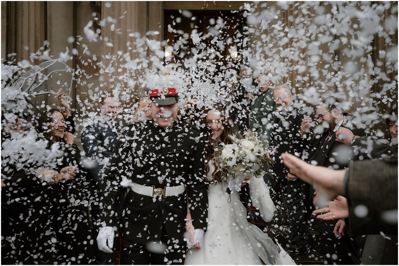 Bride and groom walking through a flurry of white confetti as guests celebrate outside the venue