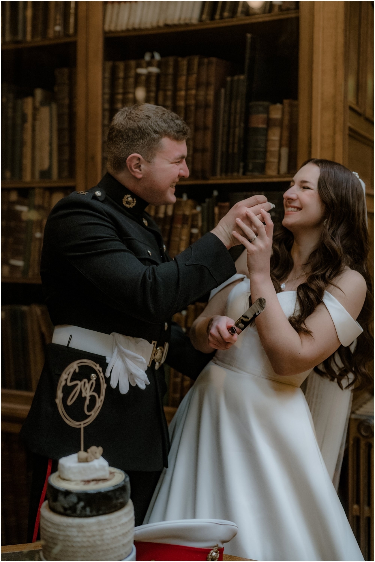Bride and groom feeding each other wedding cake and laughing in front of a wall of vintage books, with a cake topped with custom wooden initials.