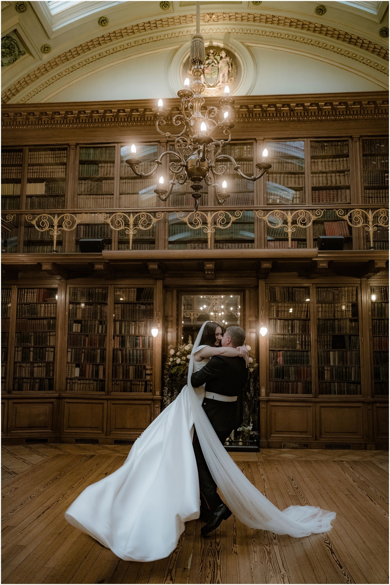 Groom lifting the bride in a dramatic embrace beneath a grand chandelier in the historic library, her dress and veil flowing across the polished wooden floor.