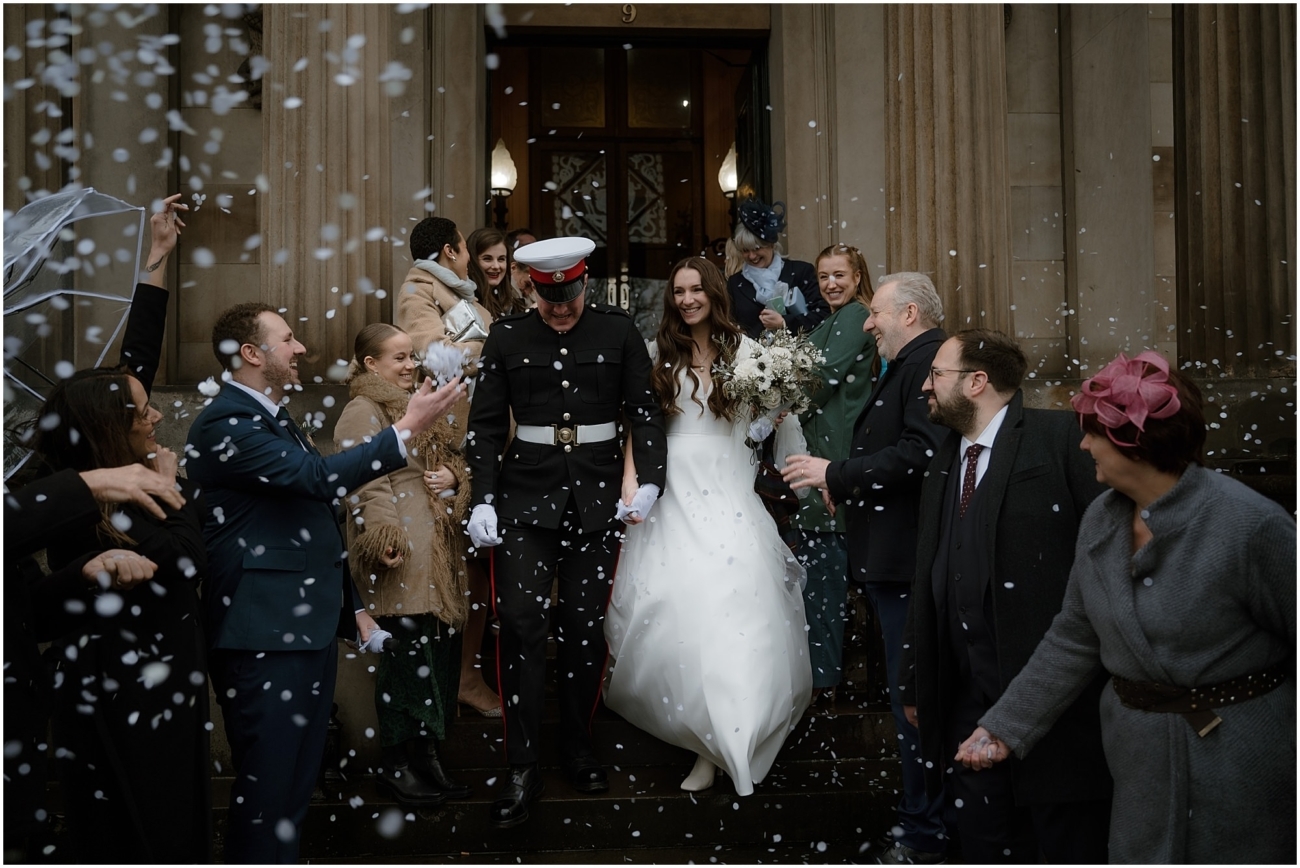Bride and groom exiting the venue, surrounded by cheering guests tossing confetti, captured mid-celebration.