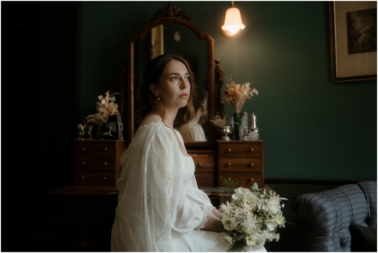 Bride in vintage-style satin gown with lace cape seated beside an antique dressing table in a Victorian flat, holding a bouquet of white wildflowers.