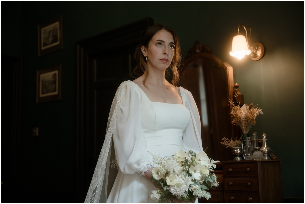 Bride standing in her vintage-style satin gown with lace cape, holding a bouquet of white wildflowers beside an antique dresser with crystal decanters in a Victorian flat.
