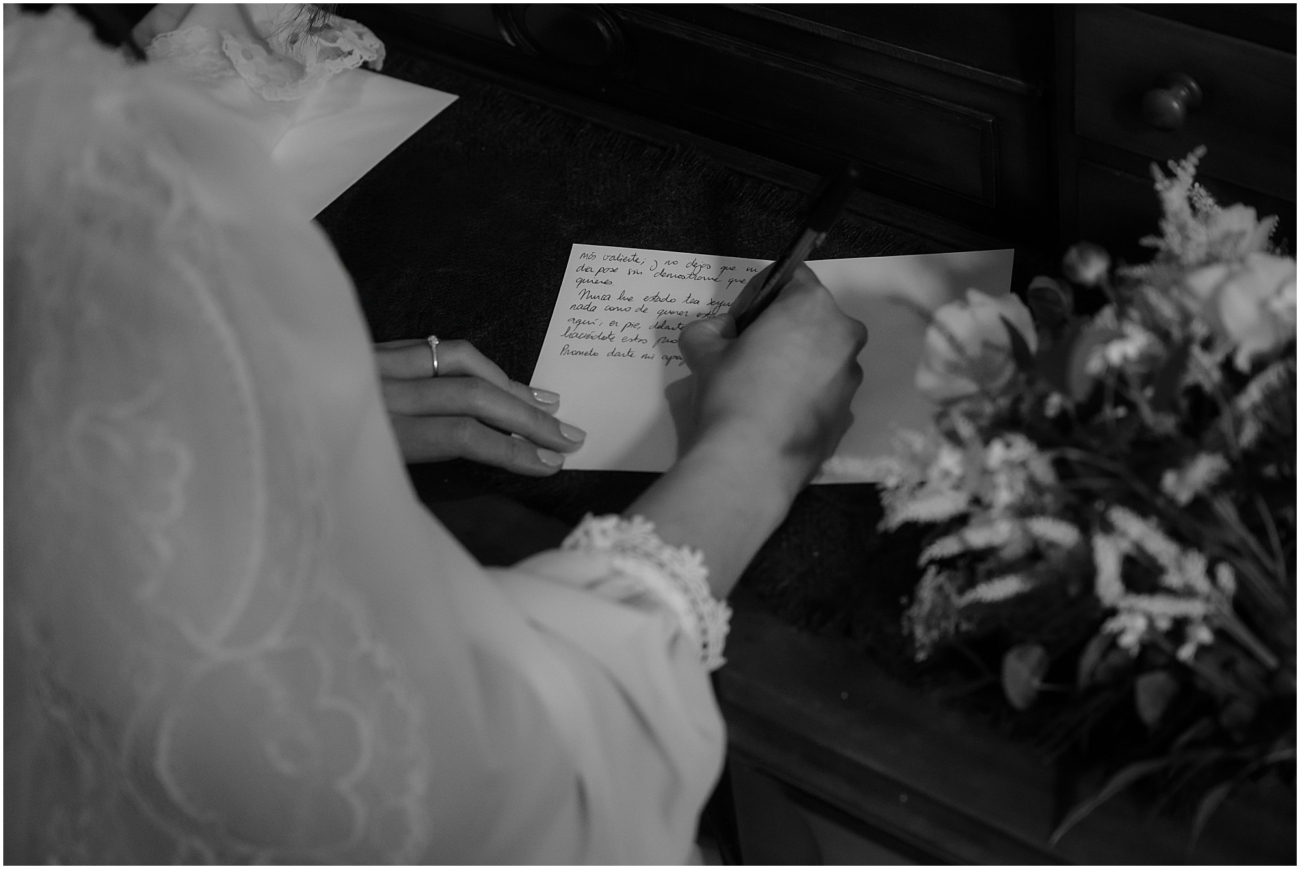 Black and white close-up of bride’s hands writing a heartfelt vows in Spanish at a dark antique desk, with her lace cape and bouquet visible.