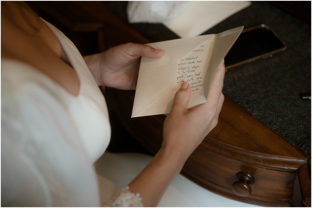 Bride holding a handwritten letter and envelope near an antique wooden desk, soft warm light highlighting the details of her gown’s lace sleeve.