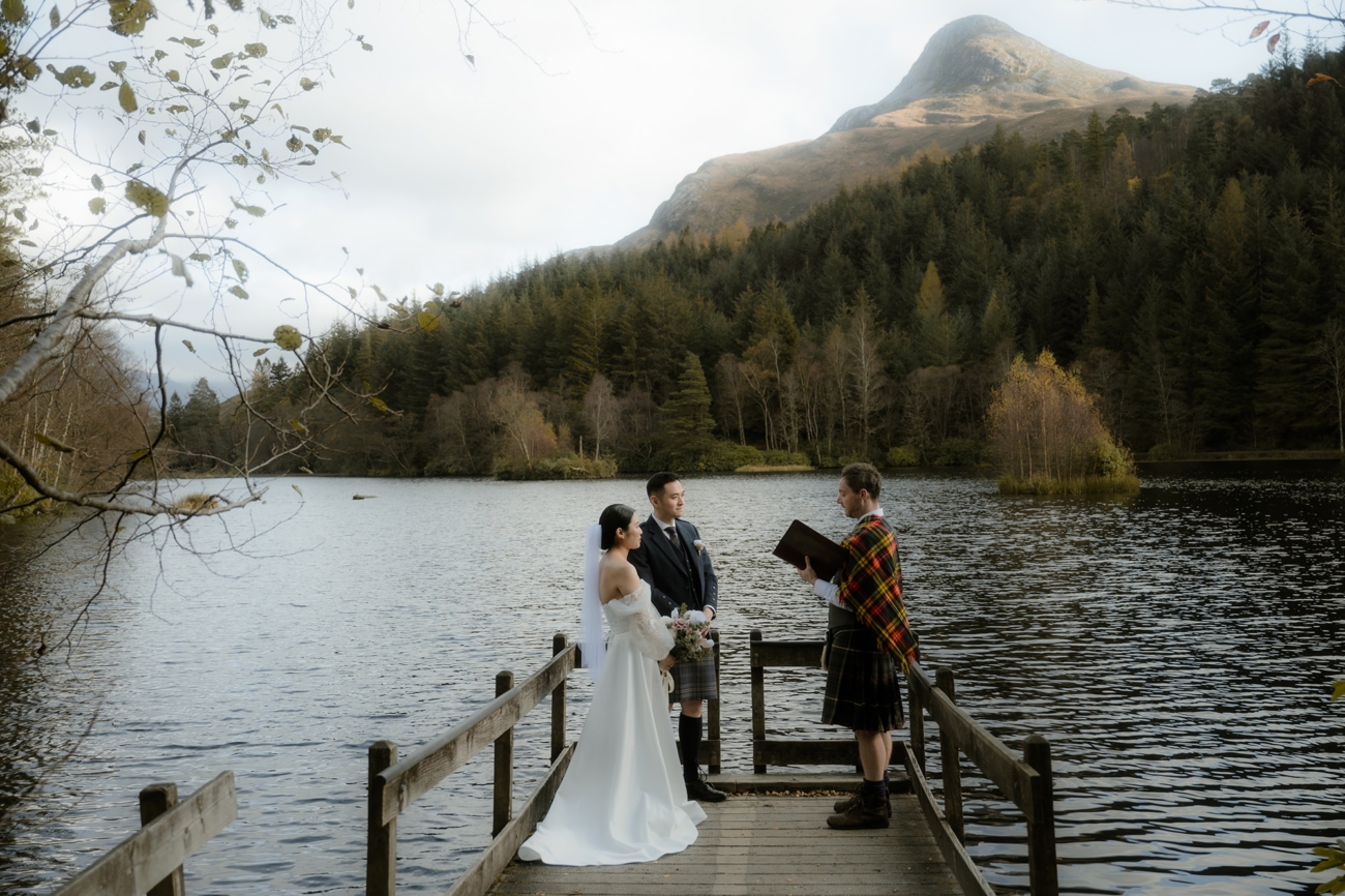 On a narrow wooden jetty over a still loch, a bride and groom face a handfasting ceremony officiant.