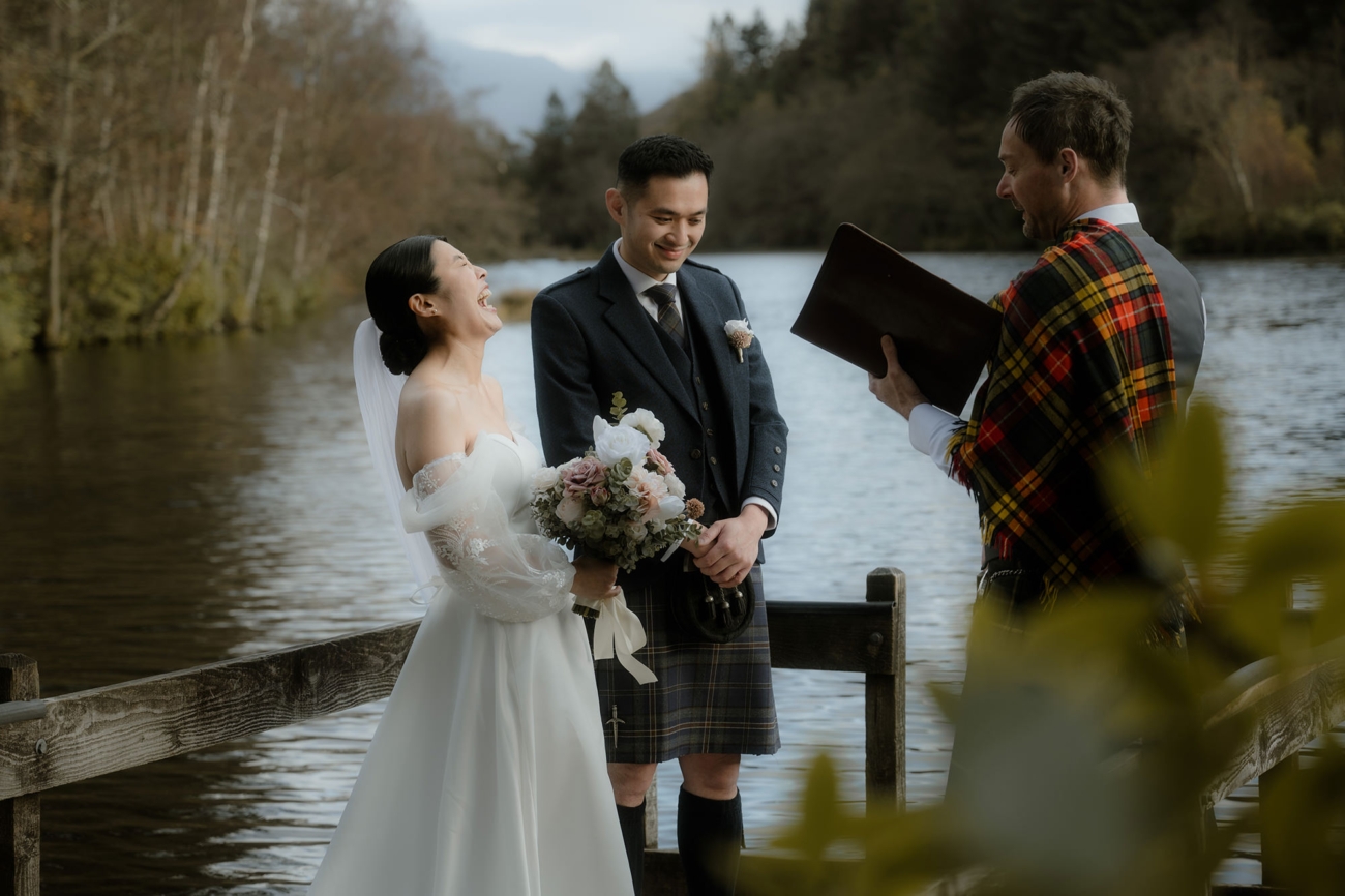A closer side view of the couple holding hands at the water’s edge. The bride laughs, clutching a bouquet of rosy and white blooms