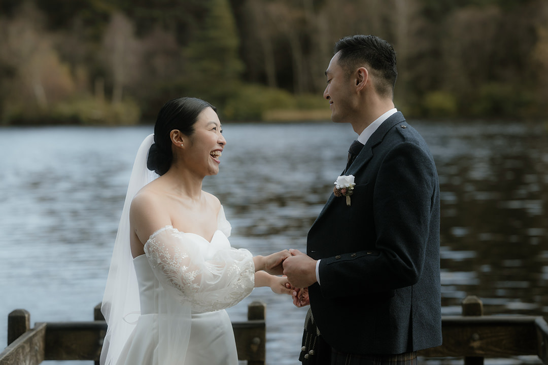 A side-profile shot of a bride and groom standing on a wooden jetty over a loch, holding hands and smiling at each other. 