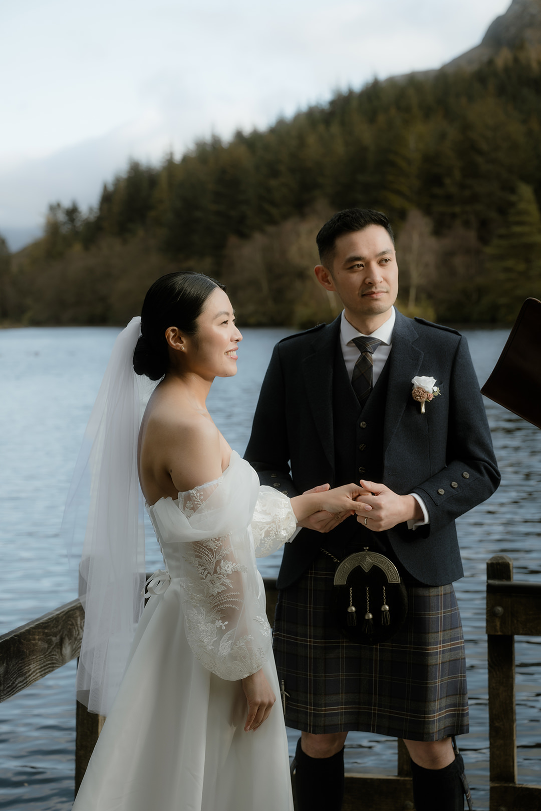 A portrait-style shot of the bride and groom standing hand-in-hand beside the loch as the celebrant speaks.