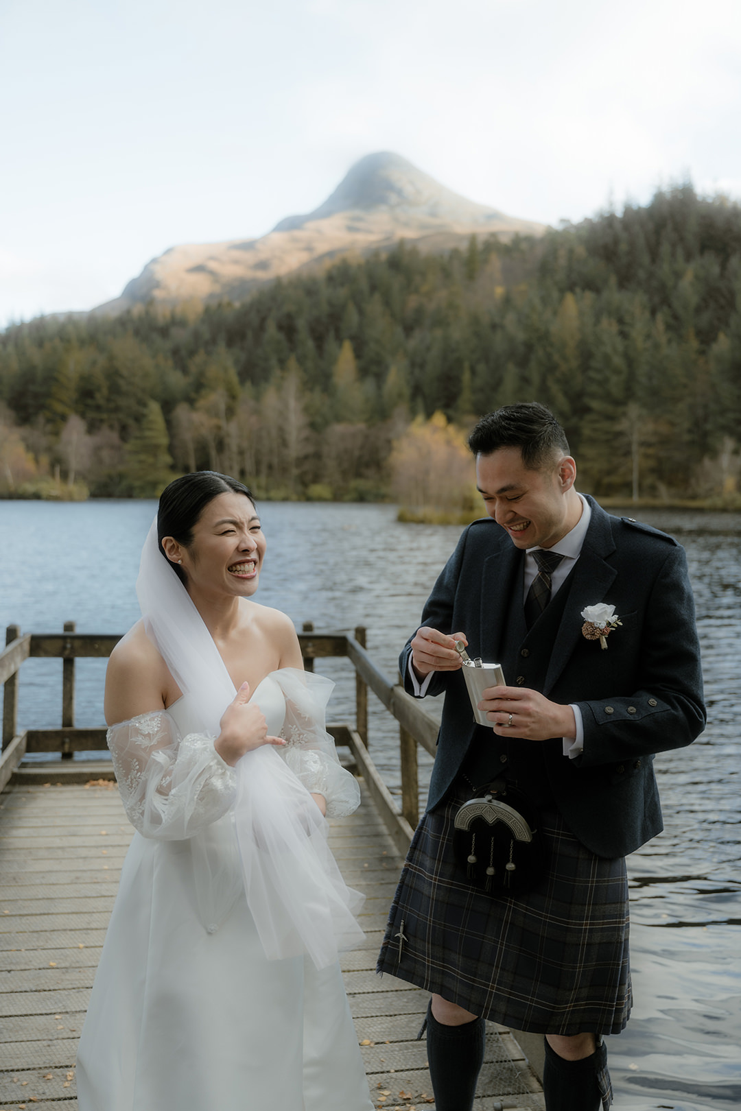 The bride and groom share a lighthearted moment after the ceremony. The bride, her veil draped over one arm, looks toward the groom with a bright smile as he holds a silver flask.