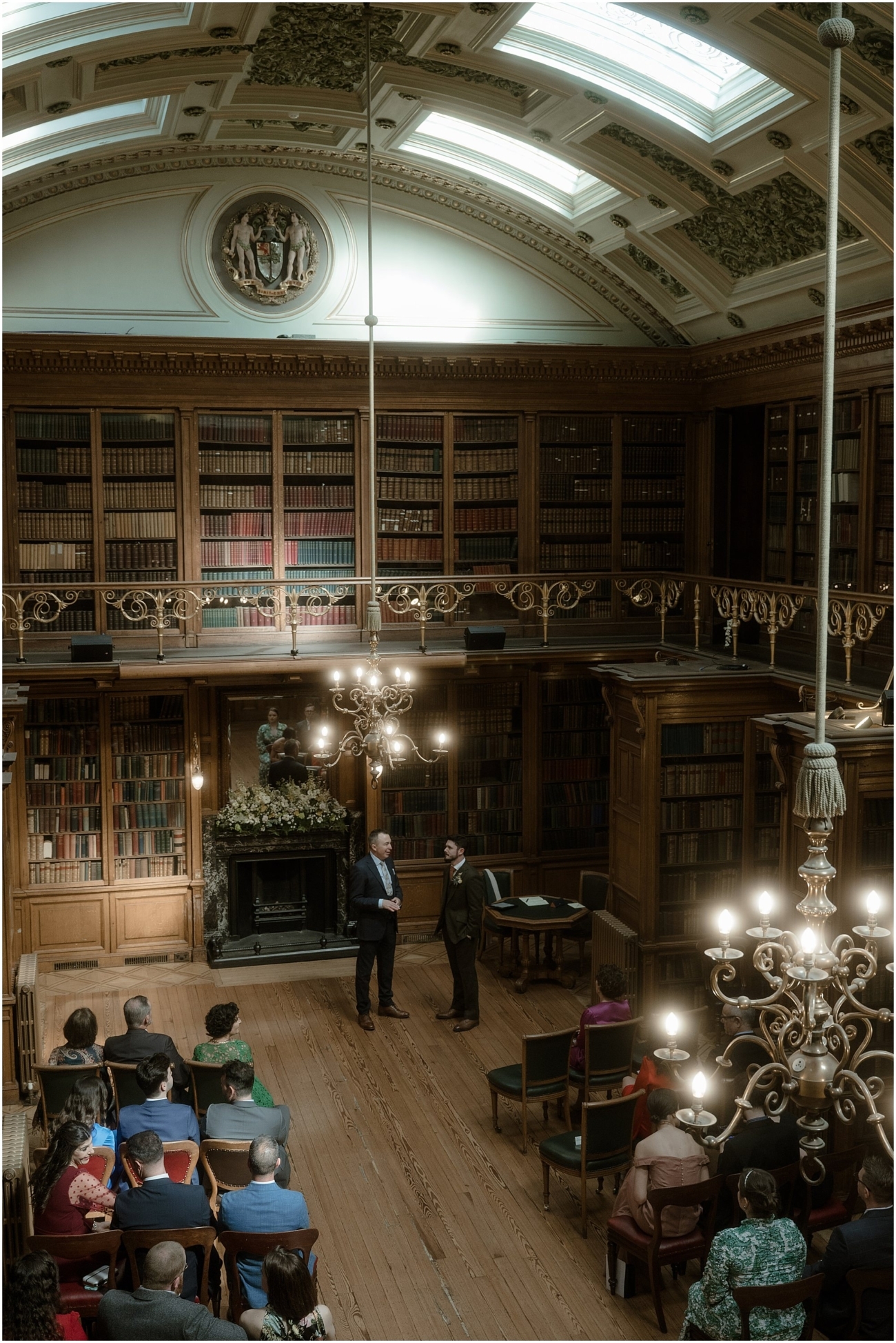 A wide-angle shot of the wedding ceremony inside a grand library with ornate vaulted ceilings, skylights, and walls lined with towering bookshelves. The couple stands at the front near a large fireplace adorned with floral arrangements. The chandelier’s warm glow fills the space as guests look on.