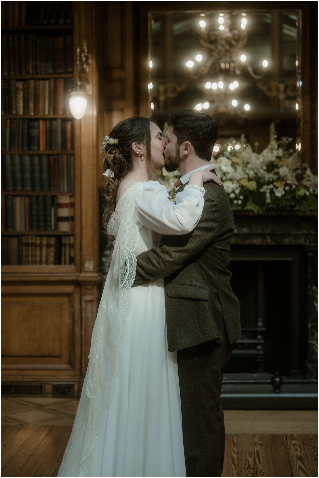 The couple share their first kiss, standing close with arms wrapped around each other in front of the grand fireplace and floral arrangements.
