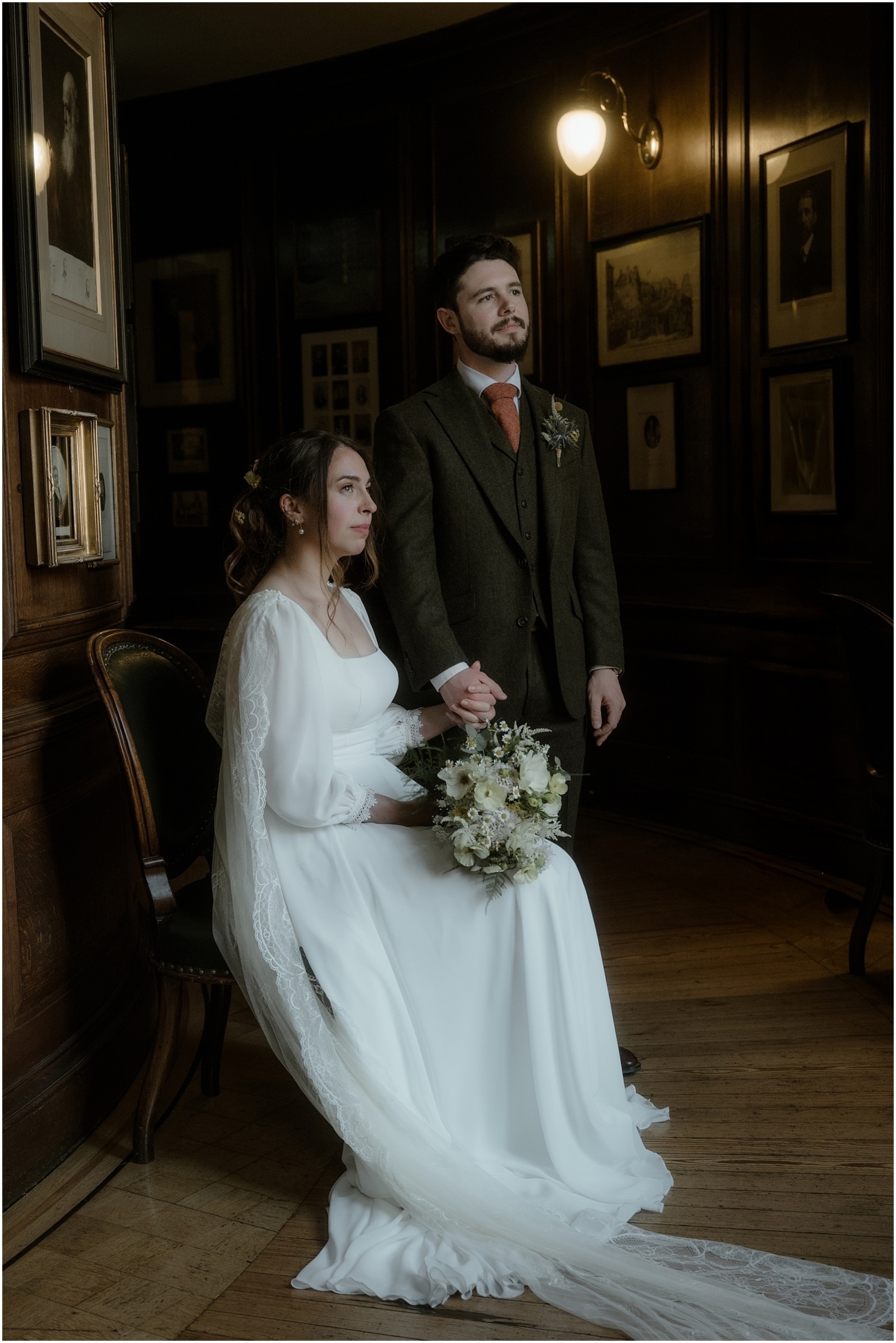 The bride sits gracefully holding her bouquet while the groom stands beside her, his hand resting lightly on hers - bathed in soft light in a richly paneled room filled with historic portraits.