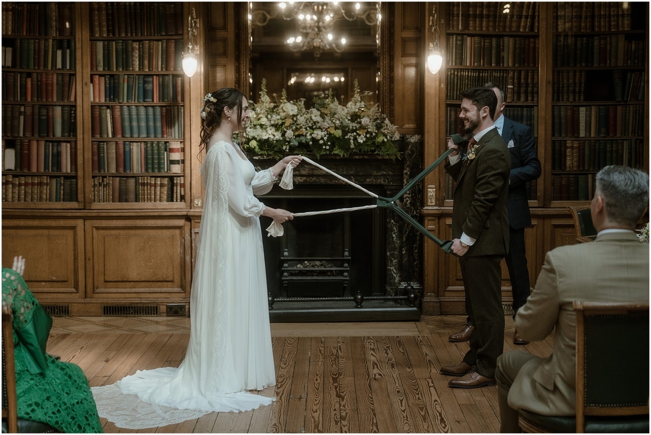 The couple stands facing each other, holding the ends of the knotted handfasting ribbons stretched between them, smiling warmly.