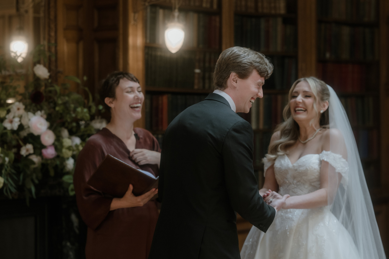 A bride and groom stand facing each other in a richly wood-panelled library lined with tall bookshelves. The bride, in an off-the-shoulder white lace gown and long veil, and the groom, in a black tuxedo and bow tie, are holding hands and laughing. To their left, a humanist celebrant in a deep burgundy dress holds an open book