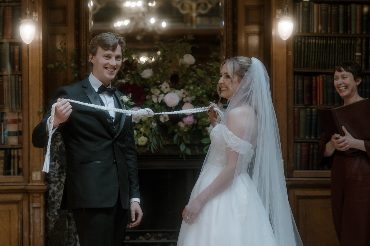 The bride and groom tie the handfasting cords during the Scottish humanist ceremony. The groom in a black tuxedo, holds one end of a braided white cord, while the bride, in her white wedding gown and veil, holds the other