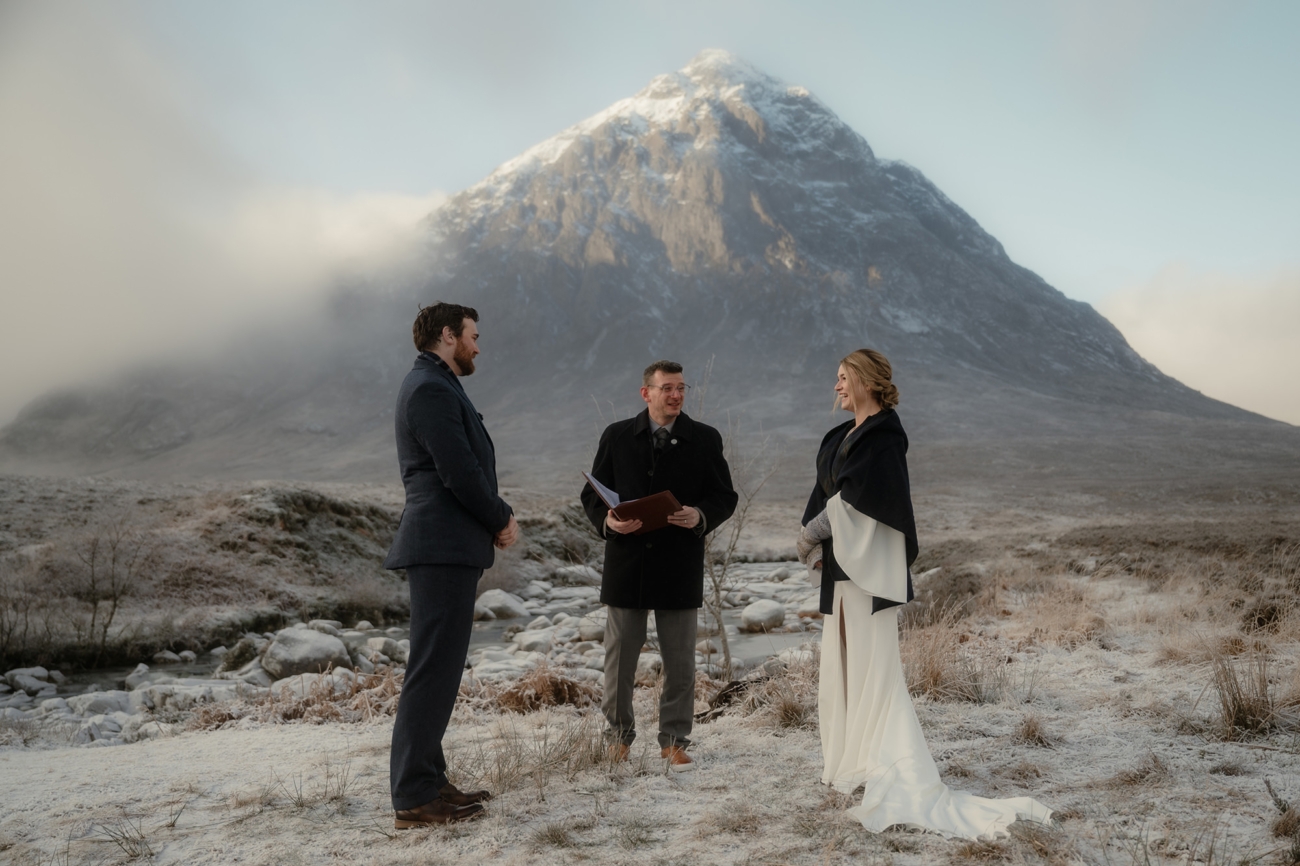 A wide‐angle view of a humanist elopement in a frost‐touched glen of Glencoe. The bride, in a sleek white dress with a dark wrap, and the groom, in a wool suit and scarf, stand facing each other on snowy grass beside a rocky stream. A celebrant holds an open book between them as a towering, snow-capped mountain looms behind under a pale winter sky.