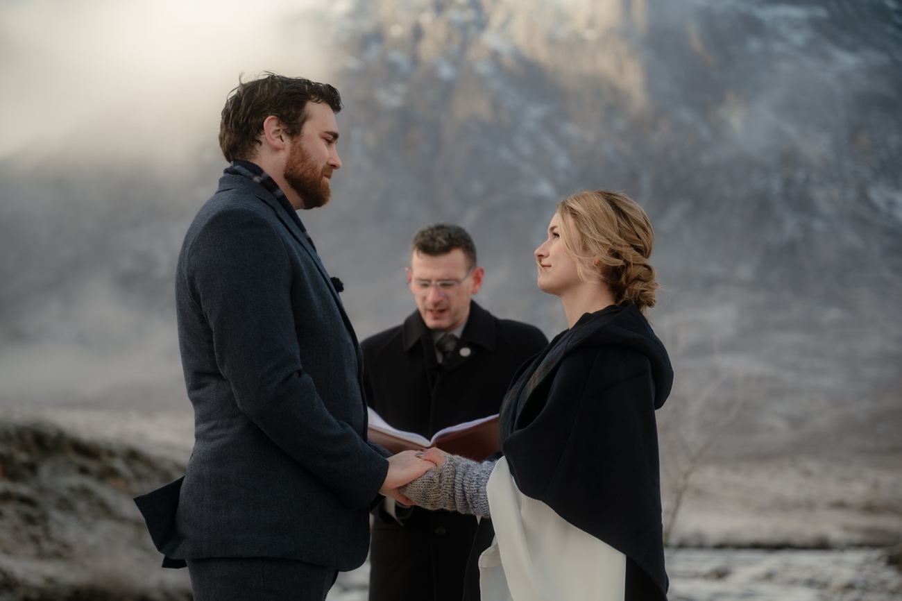 A closer, side-profile shot of the couple holding hands in front of the misty mountain while the celebrant reads from her book in soft focus behind them.