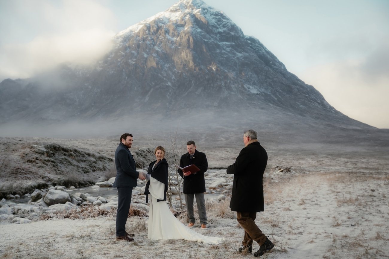 A full-scene capture showing the bride and groom clasping hands beside the celebrant, with a guest approaching as the ring bearer.