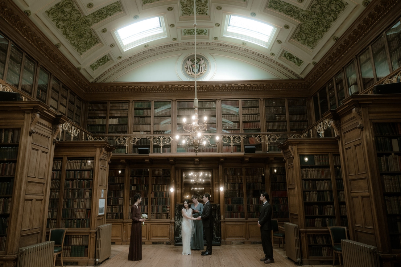 A wide view of a humanist elopement ceremony in an ornate wood-panelled library under a vaulted, skylit ceiling of Edinburgh wedding venue. The bride in a slim, beaded white gown and the groom in a dark suit stand facing each other, holding hands, as a celebrant in a muted green dress officiates before towering bookshelves and a crystal chandelier.
