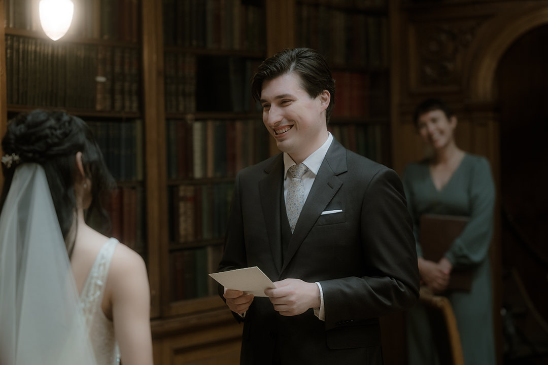 A medium shot of the groom standing at the ceremony, holding a folded card of handwritten vows and smiling warmly at his bride.